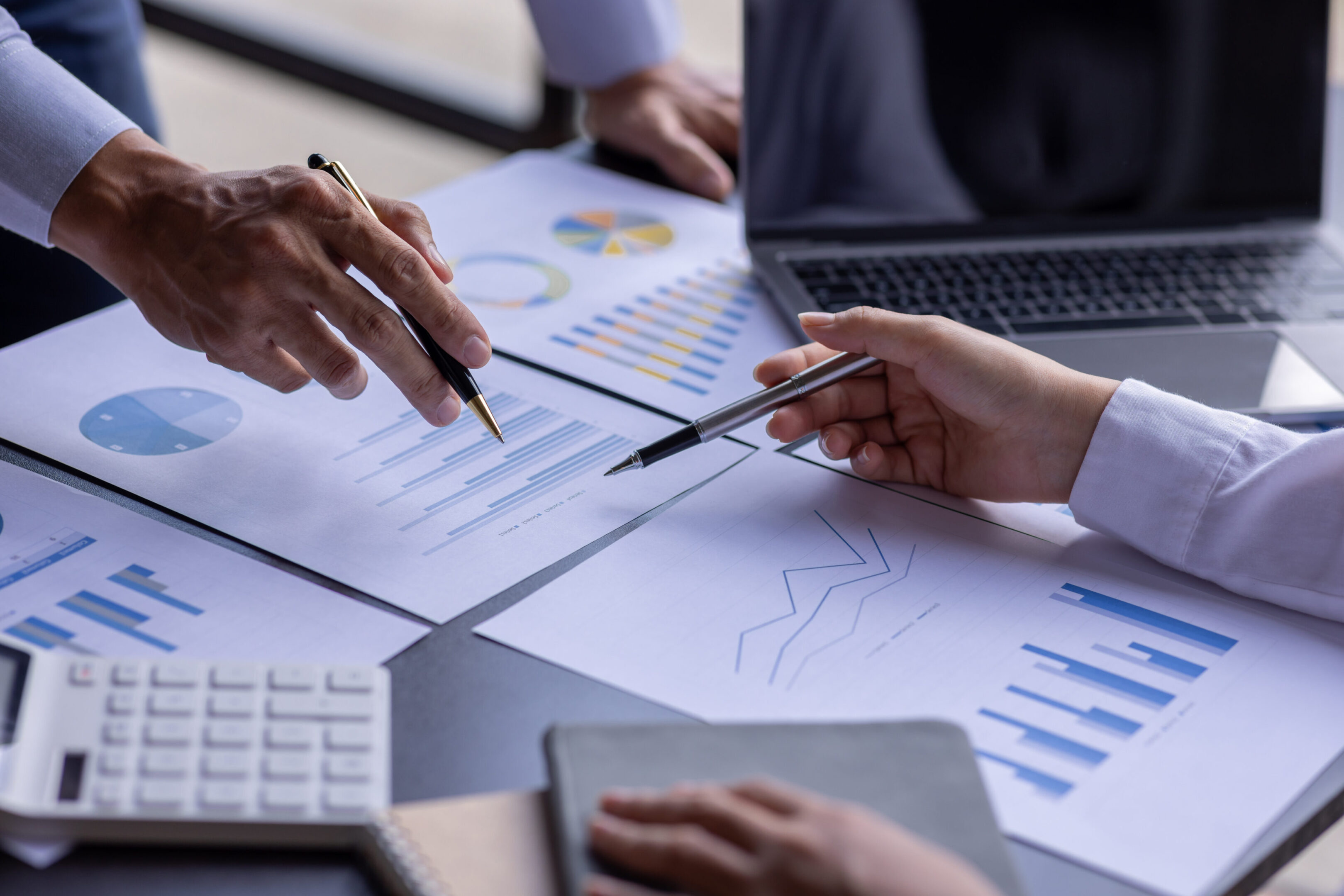 Team businesspeople or an accountant team are analyzing data charts, graphs, and a dashboard on a laptop screen in order to prepare a statistical report and discuss financial data in an office. A group of people sitting at a table with papers.